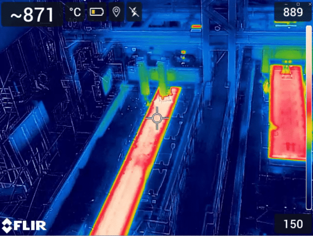 Photographie montrant la « zone formation des brames coulée continue à l’arrêt 2 » dans une aciérie ou une usine sidérurgique. On y voit une section de la ligne de coulée continue où les brames (gros blocs d’acier) sont habituellement formées, mais la production est à l’arrêt. Une image de thermographie, en fausses couleurs, affiche les températures sur la zone : les parties les plus chaudes apparaissent en rouge et jaune, tandis que les zones plus froides sont en bleu et vert. Les équipements industriels (rails, rouleaux, robinetterie) sont visibles, certains avec des points chauds signalant des résidus de chaleur, d’autres apparaissant froids. L’ensemble permet d’identifier les zones résiduelles chaudes, révélant l’état thermique de l’installation à l’arrêt